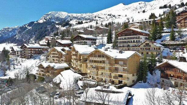 Estância de montanha coberta de neve com edifícios altos, incluindo um hotel com varandas e um teleférico visível ao fundo.