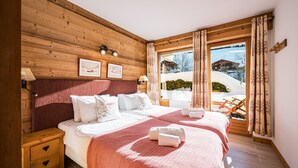 Quarto com lambris de madeira, uma cama grande com lençóis brancos, um cobertor vermelho estampado e vista para uma paisagem nevada através de uma porta de vidro.
