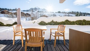 Um deck de madeira com uma mesa e cadeiras, com vista para uma paisagem nevada com casas e montanhas ao fundo.