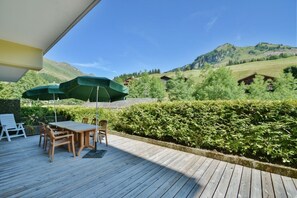 Eine Holzterrasse mit Tisch und Stühlen, einem grünen Sonnenschirm und Blick auf grüne Hügel und Häuser.