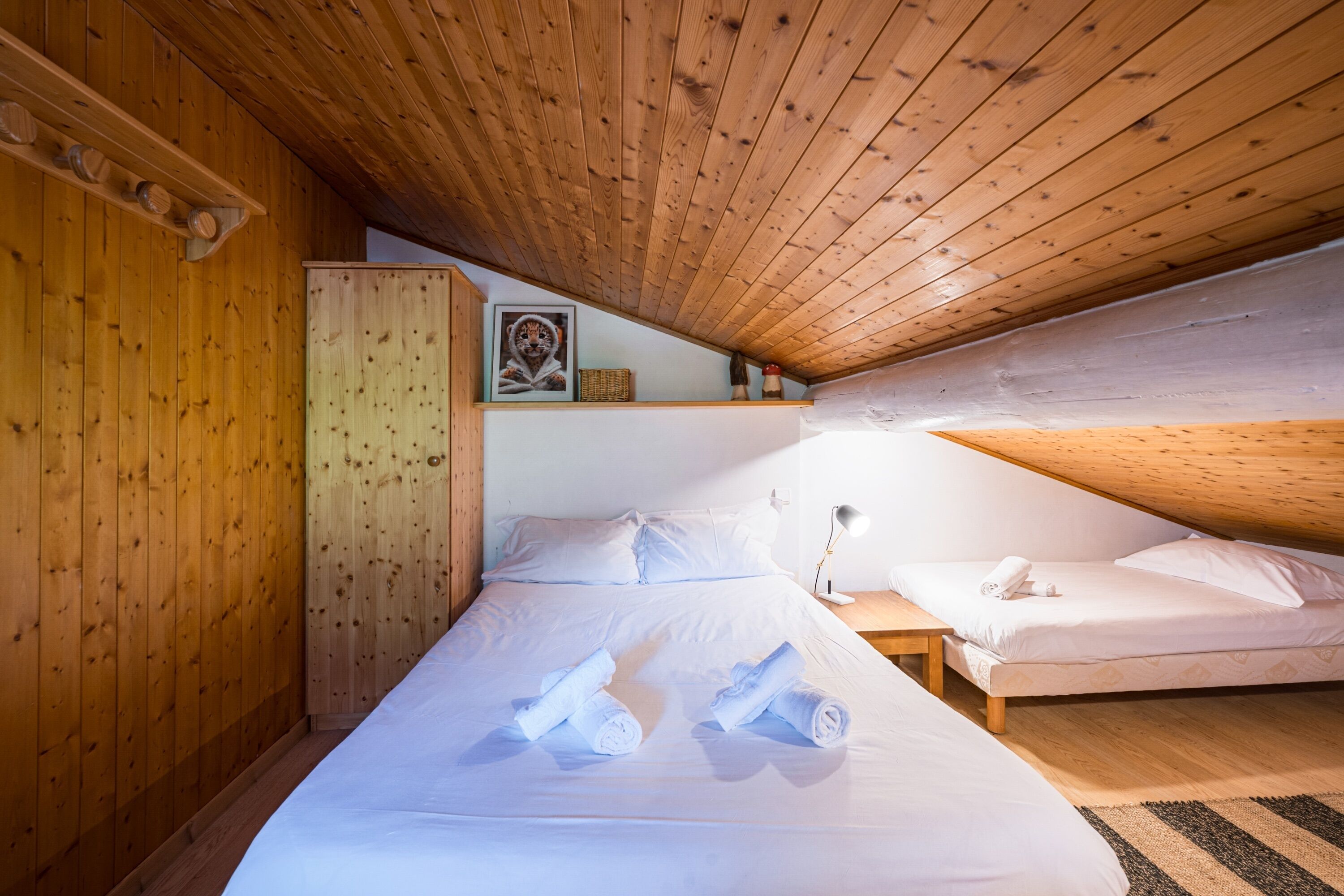 Een slaapkamer met een houten plafond, een bed met witte lakens, een nachtkastje en een plank met een ingelijst beeld.