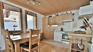 A wooden kitchen with a dining area, wooden cabinets, a stove, and a window overlooking a balcony. - Refuge Le Bardeau B2 (Le Grand Bornand)