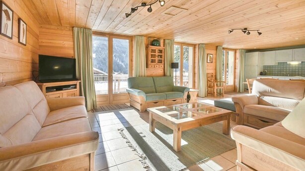 Interior de uma cabana de madeira com sofá, mesa de centro, TV e área de cozinha.