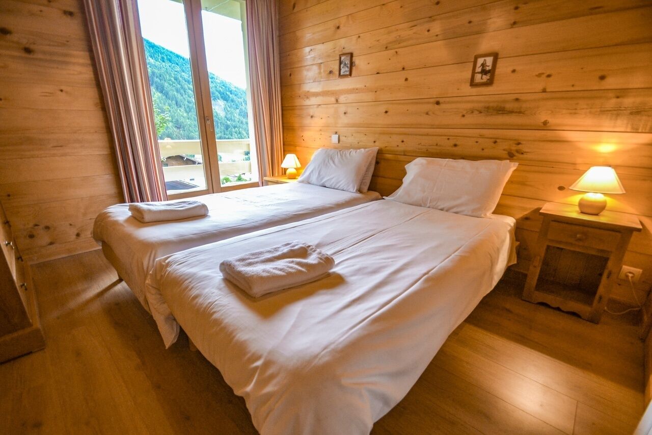Een kamer met twee bedden, houten wanden, een raam met gordijnen en een nachtkastje met een lamp.