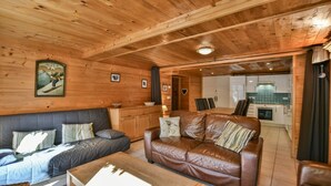 En inredning med trädetaljer, en soffa och en läderbäddsoffa, ett kök i bakgrunden och en tavla med ett bergslandskap på väggen.