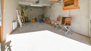 Un garaje espacioso con paredes de concreto, techo de madera y una amplia mesa blanca en el centro. Hay dos sillas plegables y una pila de leña junto a la pared.