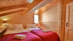 Un dormitorio de cabaña de madera con dos camas, cada una con ropa de cama roja y almohadas blancas. Hay una ventana que proporciona luz natural y una puerta a la derecha.