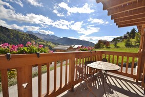 A wooden balcony with a table and chairs, overlooking a mountainous landscape with houses and greenery.