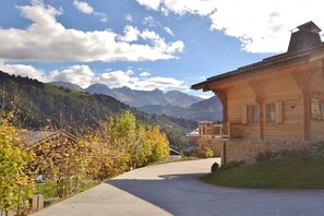 A wooden house with a balcony, surrounded by a gravel driveway and autumn foliage.