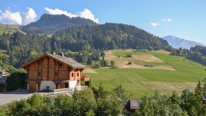 Un chalet in legno con un balcone, sullo sfondo di campi verdi e una montagna boscosa.