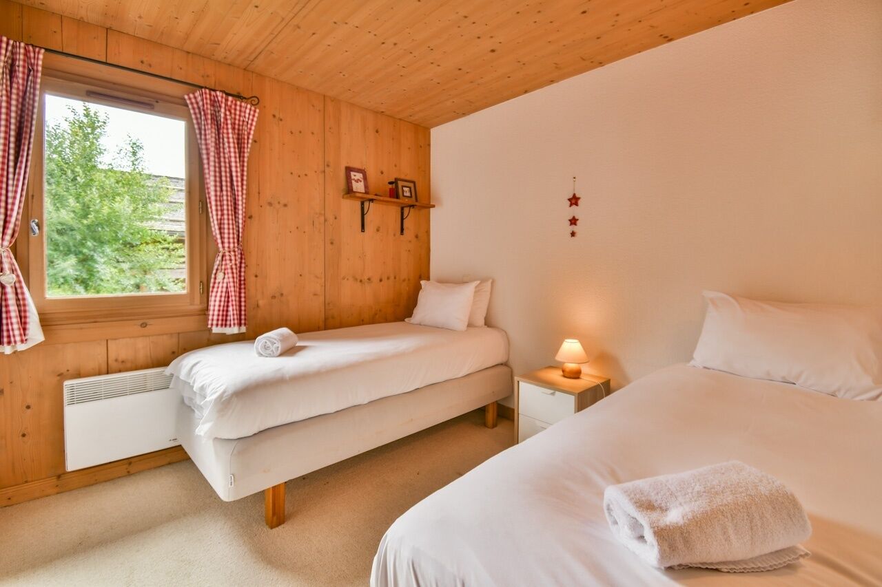 A bedroom with two beds, a window with checkered curtains, a bedside table with a lamp, and a wall-mounted shelf with pictures.