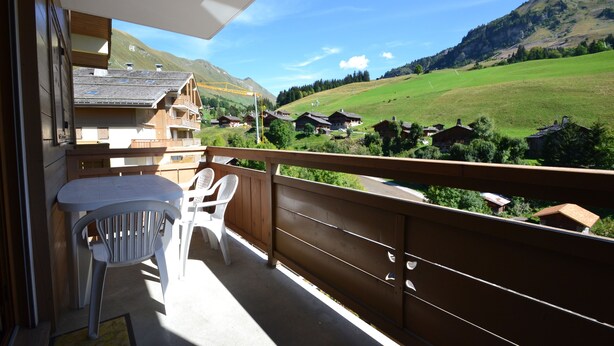 A balcony with a table and chairs, overlooking a village with houses and a mountain.