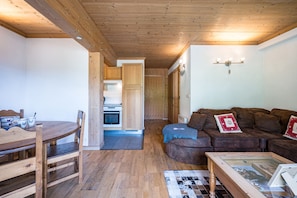 Un salón acogedor con techo de madera, un sofá marrón, una mesa de comedor con sillas y una cocina visible a través de una puerta abierta.