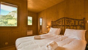 A wooden cabin bedroom with a bed, two nightstands, a lamp, and a view of a green landscape through a window. - Refuge Le Bardeau B4 (Le Grand Bornand)