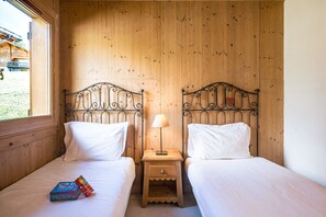 A room with two beds, each with a wooden headboard and white bedding. There is a small wooden nightstand with a lamp between the beds. A board game is visible on the floor beside one bed. - Refuge Le Bardeau B4 (Le Grand Bornand)