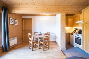 A wooden interior with a dining table, chairs, a kitchen area, and a door. - Refuge Le Bardeau B4 (Le Grand Bornand)