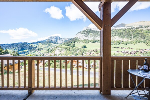 Un balcón de madera con vistas a un paisaje montañoso, una mesa con una botella y un vaso, y una silla.