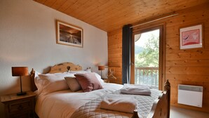 A wooden-paneled bedroom with a bed, bedside tables, and a window with a view of greenery. - Chalet Mitoyen - Rosa (Le Grand Bornand)