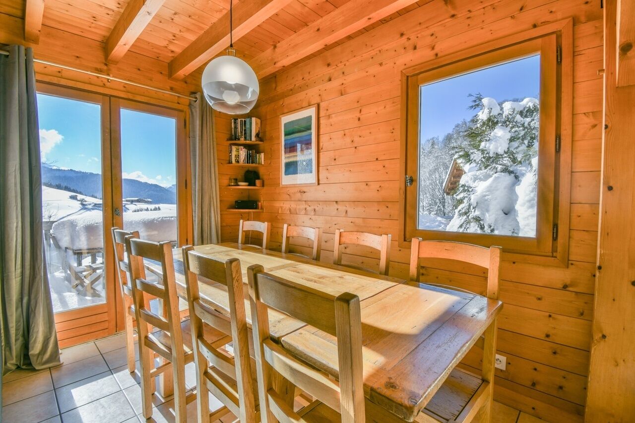 A wooden cabin with a dining table and chairs, large windows with snowy views, and a hanging light fixture.