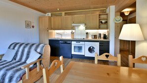 Una cocina compacta con armarios de madera, lavavajillas, horno y lavadora. Hay una mesa de comedor de madera con sillas y un sofá con una manta a rayas.