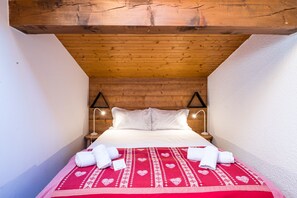 A small, cozy room with a wooden ceiling, a bed with white linens, two bedside lamps, and a red patterned bedspread. - Mont Blanc 4 - Three Bedroom Apartment, Sleeps 6 (Le Grand Bornand)