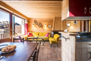 A cozy cabin interior with a wooden dining table, yellow sofa, and a kitchen area with a red cabinet. - Mont Blanc 4 - Three Bedroom Apartment, Sleeps 6 (Le Grand Bornand)