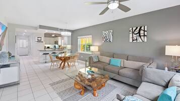A modern living room with a glass coffee table, grey sofa, and a dining area with wooden chairs.