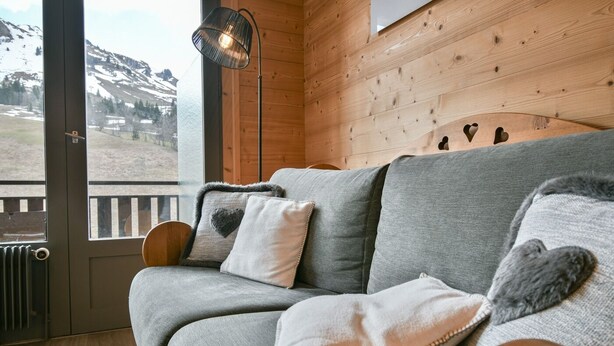Interior acolhedor de cabana com sofá cinzento, almofadas em formato de coração e vista para uma paisagem nevada através de uma grande janela.