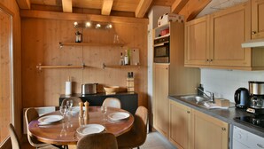 A wooden interior with a dining table set for four, a kitchen area with wooden cabinets, and a built-in oven. - Refuge Le Gypaète G6 (Le Grand Bornand)