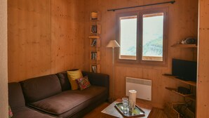 A cozy wooden cabin interior with a brown leather sofa, a small table with glasses and a tray, a lamp, and a television on a shelf. - Refuge Le Gypaète G6 (Le Grand Bornand)