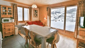 A cozy wooden cabin with a dining table, chairs, a sofa, and a TV. Large windows offer a view of a snowy mountain landscape. - Refuge Le Bardeau B1 (Le Grand Bornand)