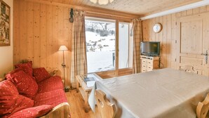 A cozy room with a wooden ceiling, a bed, a sofa, a TV, and a window with a view of a snowy landscape. - Refuge Le Bardeau B1 (Le Grand Bornand)