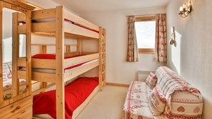 A bunk bed room with a red bedspread, a sofa with patterned cushions, a window with curtains, and a wall-mounted light fixture. - Refuge Le Bardeau B1 (Le Grand Bornand)