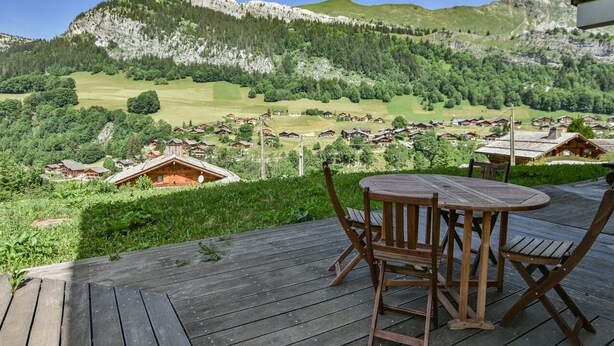 A wooden deck with a table and chairs overlooking a village with houses and mountains in the background. - Refuge Le Bardeau B1 (Le Grand Bornand)