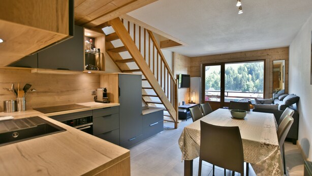 Cocina moderna con armarios de madera, estufa empotrada y comedor con mesa y sillas. Una escalera de madera conduce a un dormitorio en el altillo.