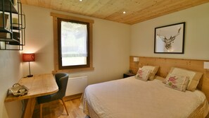 A bedroom with a wooden headboard, a bed with floral pillows, a desk with a lamp, and a window with a view of greenery.