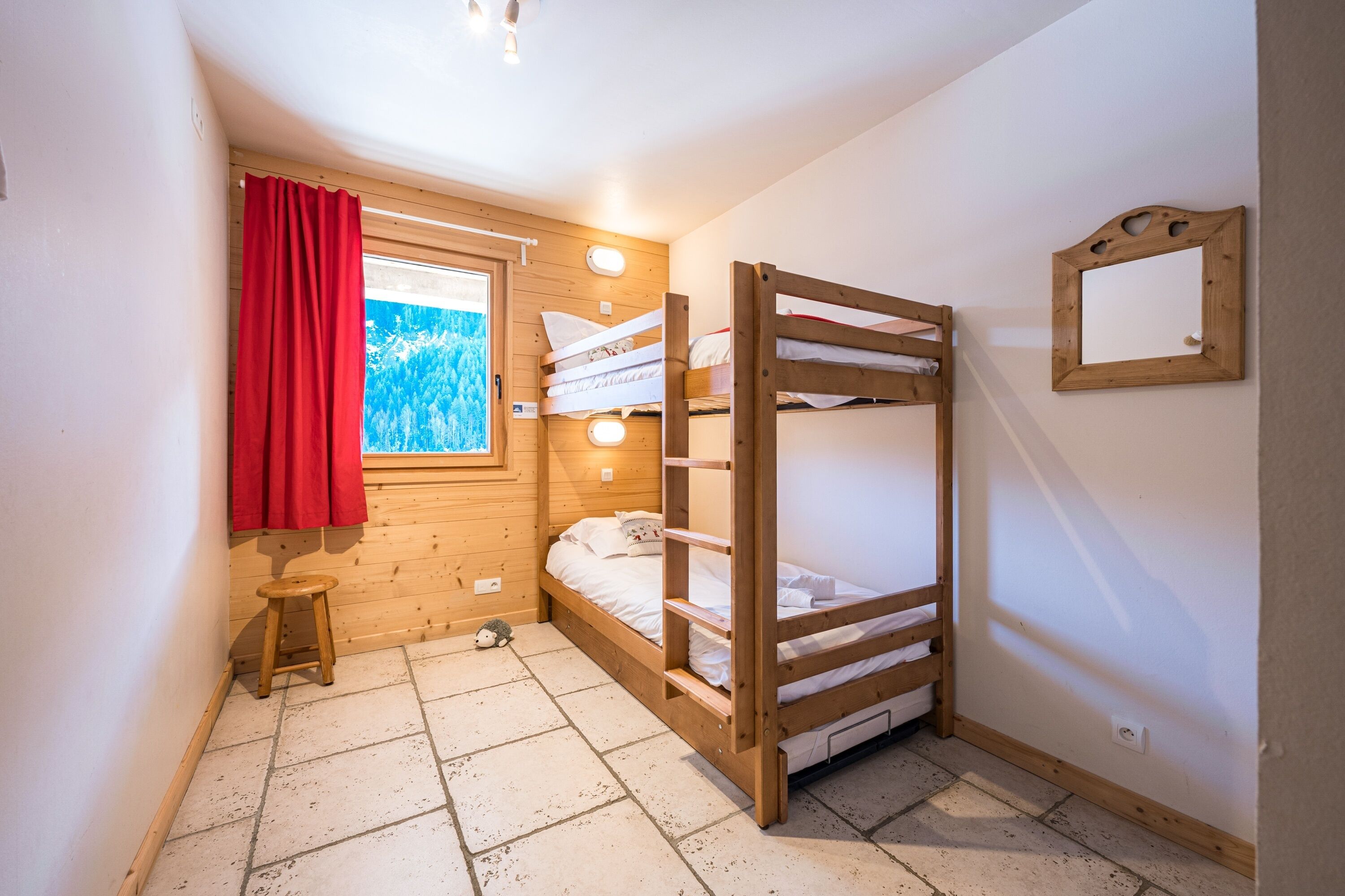 Une petite pièce accueillante, avec un lit superposé, un tabouret en bois, une fenêtre ornée d’un rideau rouge et un miroir accroché au mur.