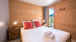 Une chambre aux murs lambrissés, avec un lit, deux tables de chevet et une vue sur les montagnes par une porte vitrée.