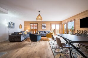 A spacious living area with a sofa set, a dining table, and a large window offering a view of the outdoors.