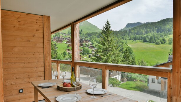 Mesa de madera dispuesta con platos, copas de vino y una botella, con vistas a un paisaje montañoso pintoresco.