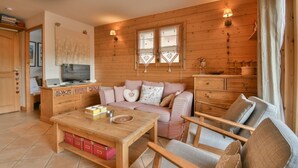 A cozy living room with a sofa, a wooden coffee table, and a TV. There are wooden walls and a window with curtains.