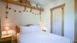 A bedroom with a wooden bed, white bedding, a nightstand with a lamp, and a shelf with decorative items.