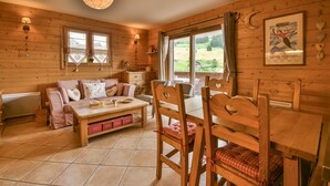 A cozy wooden cabin interior with a dining table, chairs, a sofa, and a large window offering a view of the outdoors.