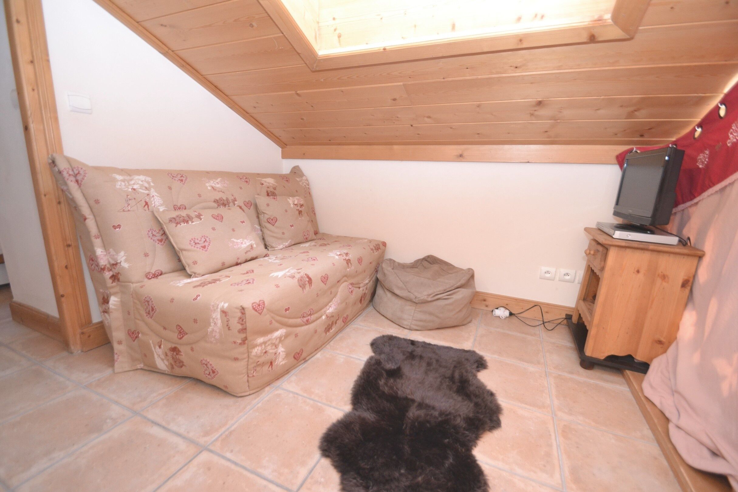 Une pièce chaleureuse avec un plafond en pente, un lit recouvert d’une literie à motifs floraux, une petite armoire en bois avec un téléviseur, et un chien noir allongé sur le sol.