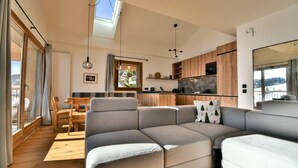Una sala de estar moderna con un sofá modular, un comedor con mesa de madera y una cocina con gabinetes de madera y un ventanal grande.