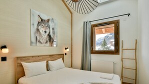 Un dormitorio con cama de madera, ventana con vista a montañas nevadas, cuadro colgado en la pared con un lobo, y una escalera de madera apoyada contra la pared.