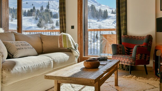 Ein gemütliches Wohnzimmer mit einer Couch, einem Holztisch und einem farbenfrohen Sessel, mit einem großen Fenster und Blick auf einen verschneiten Berg.