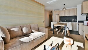Ein modernes Wohnzimmer mit Sofa, Esstisch und Stühlen. Es gibt eine Küchenzeile mit Holzausbau und Arbeitsplatte.