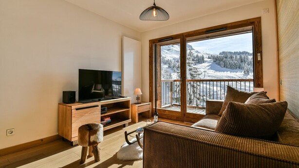 Ein gemütliches Wohnzimmer mit Fernseher, Holzmöbeln, einem Hund und einem Balkon mit Blick auf einen verschneiten Berg.