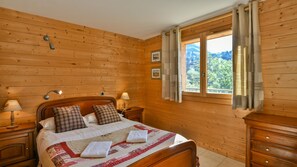 Un dormitorio con paneles de madera, una cama, dos lámparas de noche, un ventanal con cortinas y vistas al verdor.